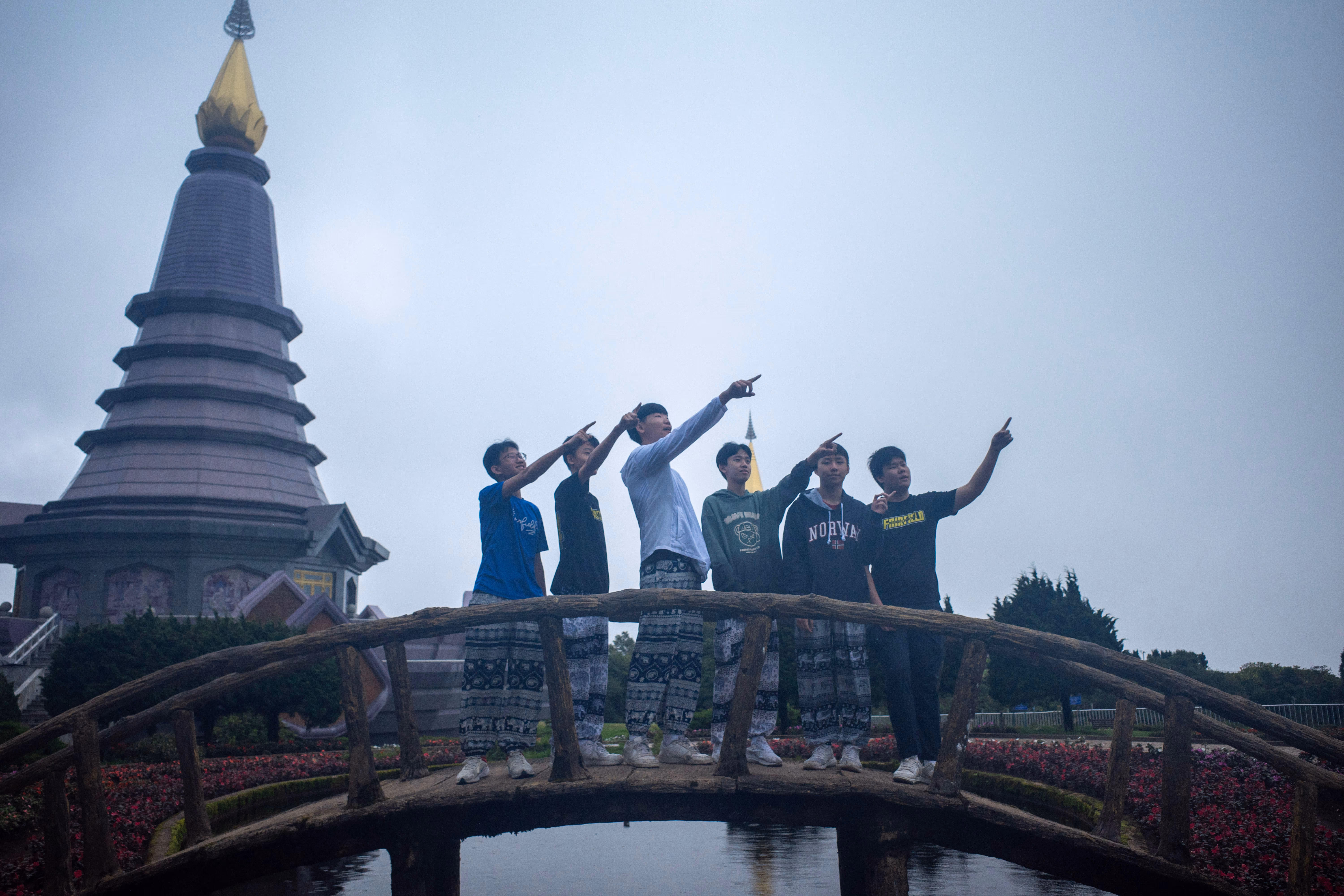 ix Secondary 3 students on a Trips for International Experience (TIE) journey pose together on a rustic, arched wooden footbridge over a small pond in Thailand. The students are lined up side-by-side, each smiling and pointing their right arm upward toward the sky in a synchronized gesture. Most of them are wearing loose, black-and-white patterned traditional Thai elephant pants paired with t-shirts and hoodies. In the background, a large, multi-tiered dark grey pagoda with a prominent gold spire rises against a misty, overcast sky, surrounded by landscaped flowerbeds.
